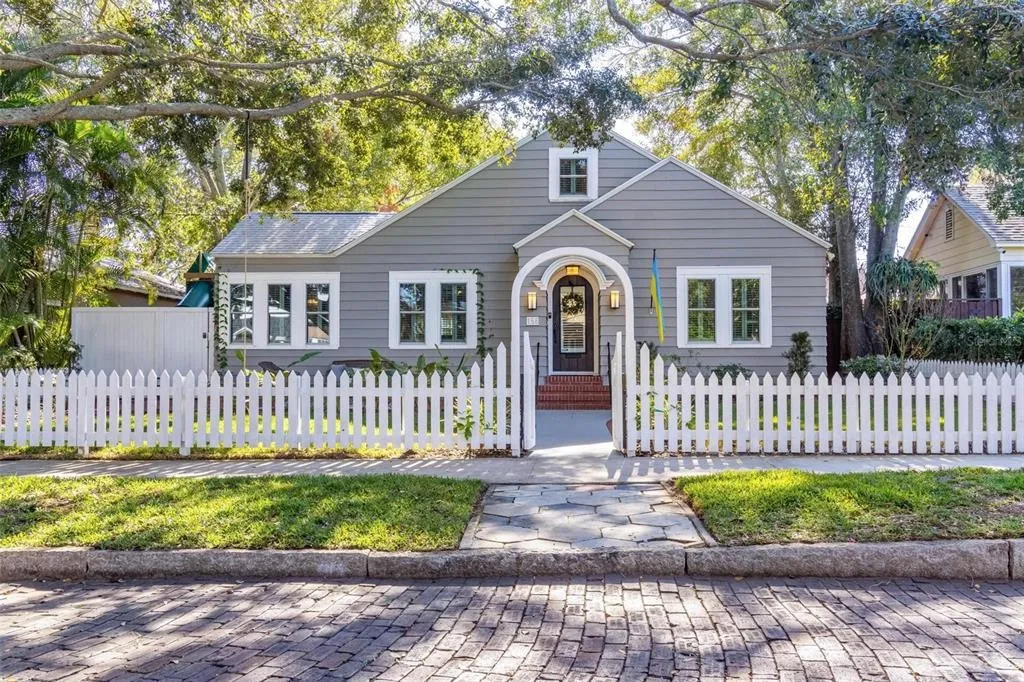 Exterior view of small gray home with white window trim and white picket fence in front. Surrounded by tall trees.
